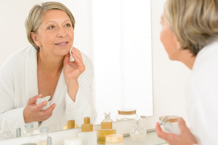 Pretty senior woman in bathroom looking at herself in mirror while applying moisturizing lotion