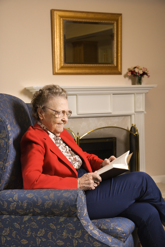 Elderly Caucasian woman reading book in chair at retirement community center.