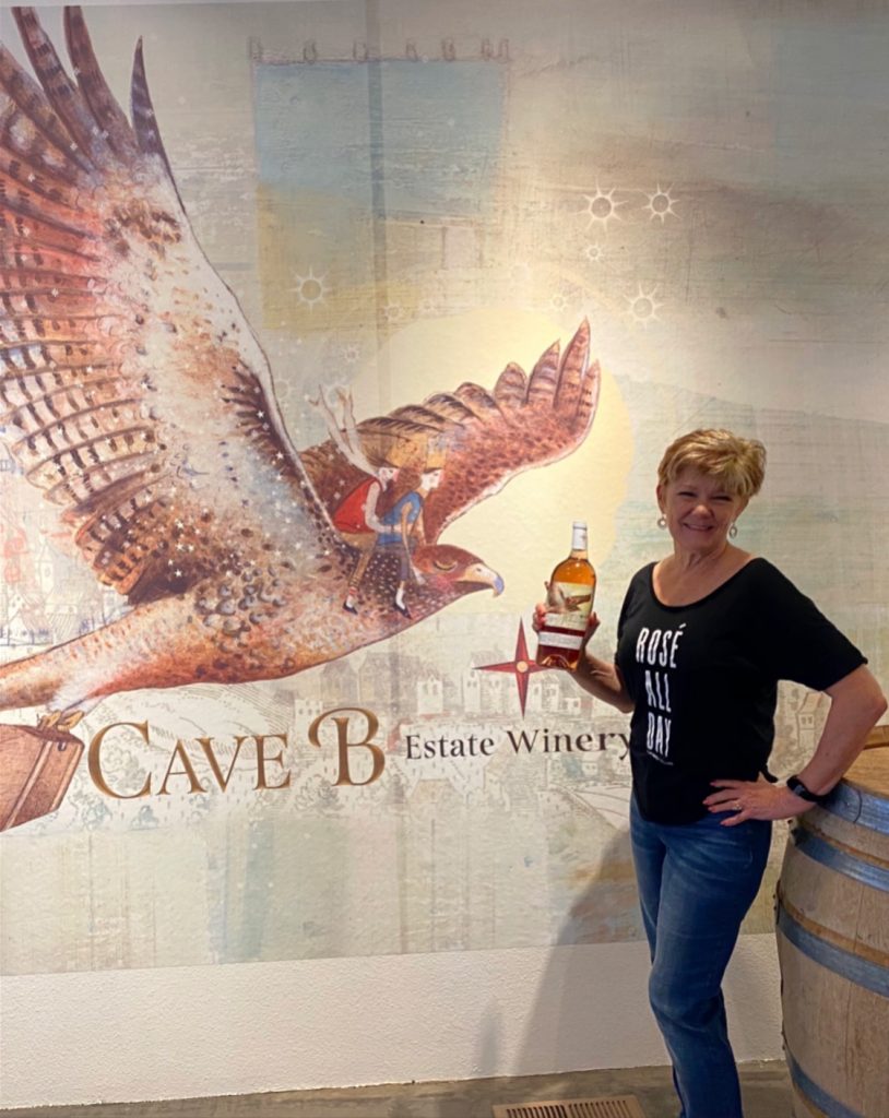 Shelley standing in front of the Cave B label mural and holding a bottle of rose'