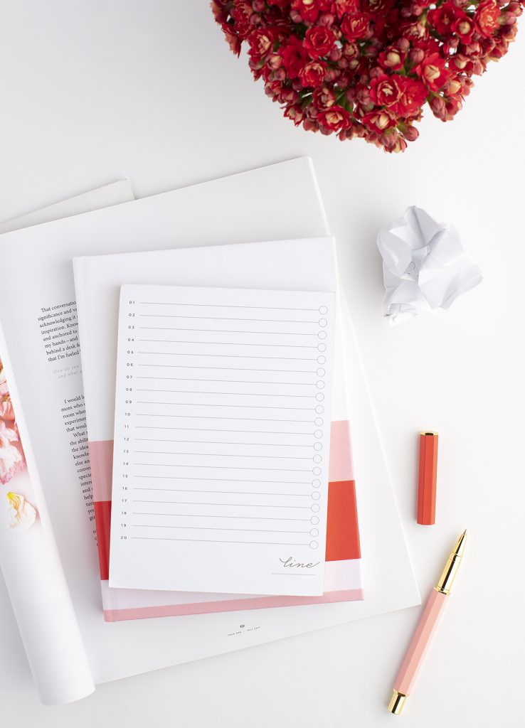 notepad on top of a journal with a pen beside it. red flowers in the background