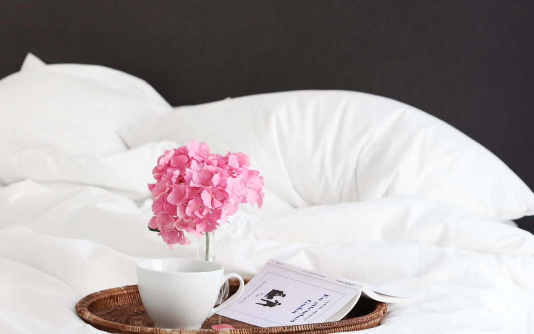 a bed tray on an unmade bed. On the tray is coffee, a book and a pink flower
