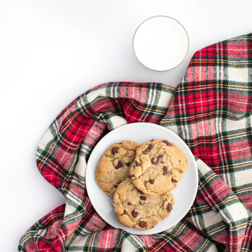 a plate of 3 chocolate chip cookies lying on a red plaid blanket with a cup of milk on the side