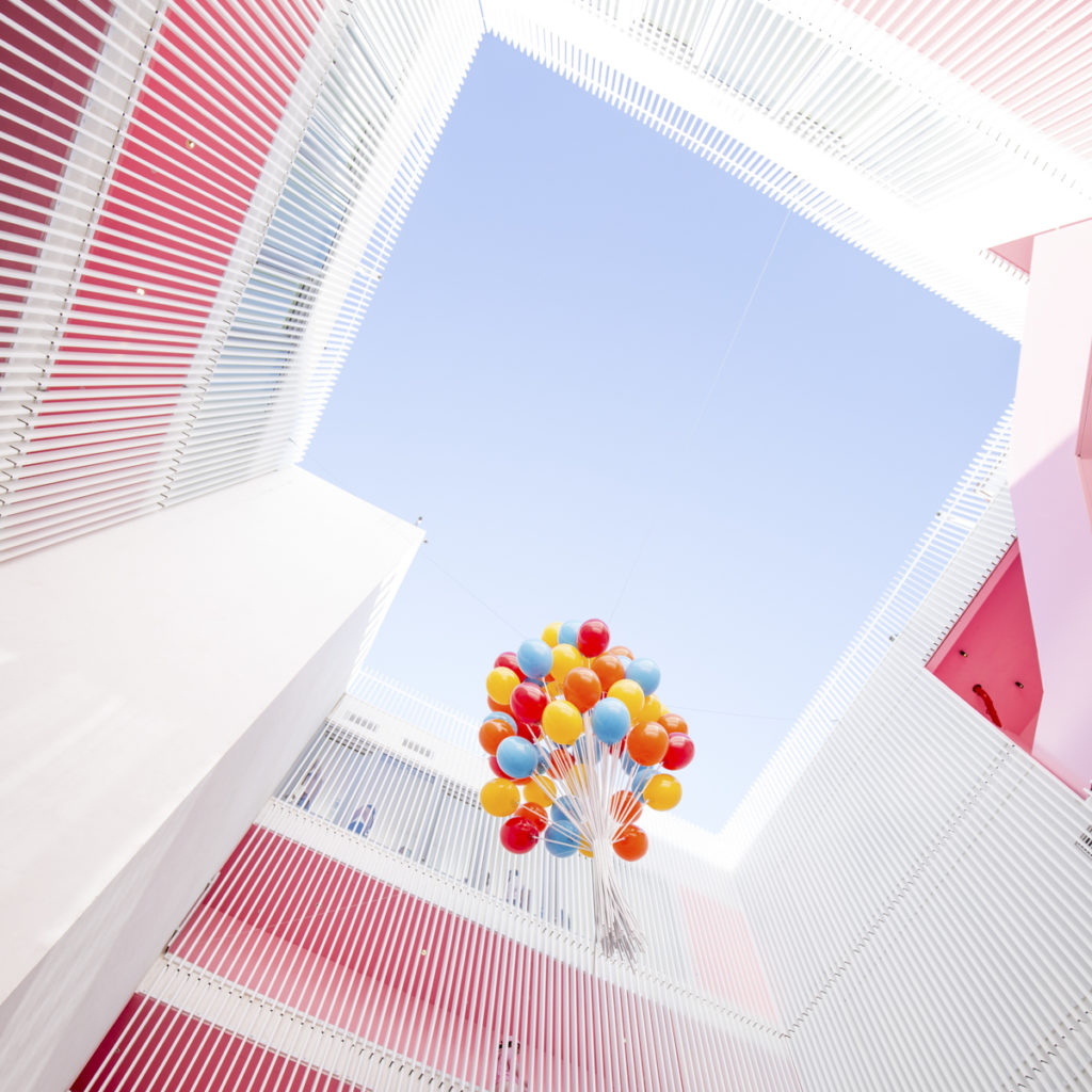 Alarge bunch of helium balloons floating upward through the balcony of a pink building.