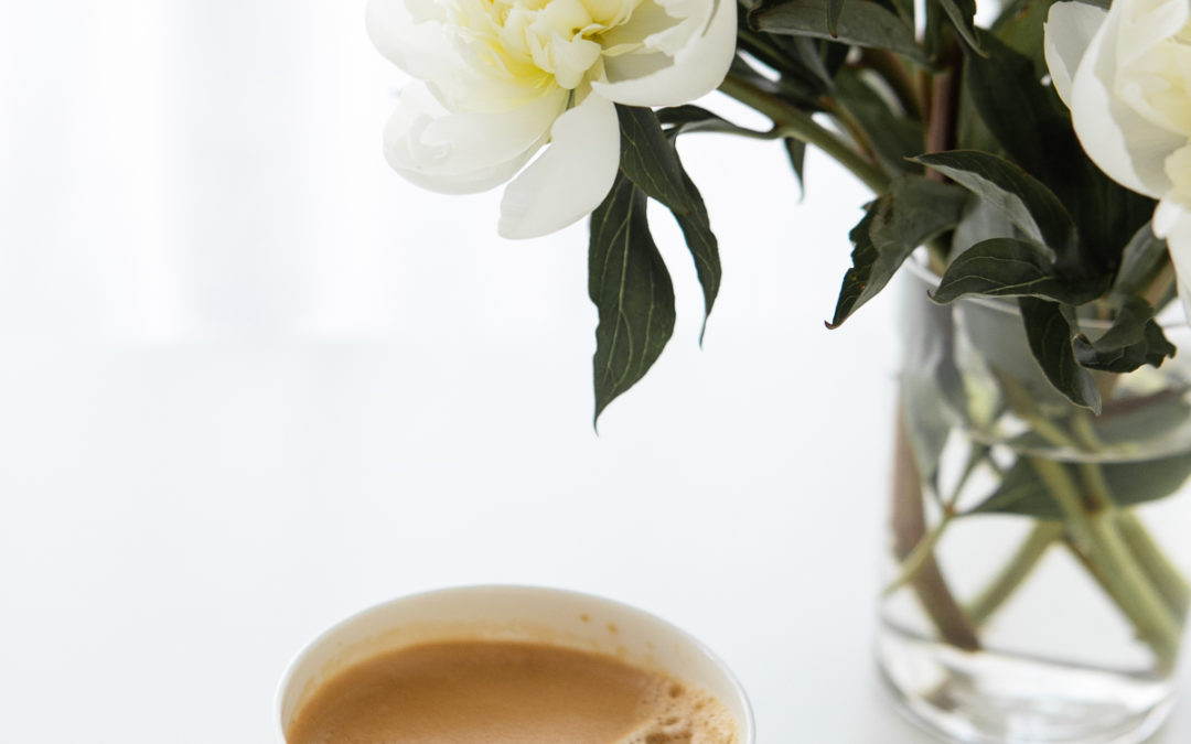 A cup of coffee with cream next to a vase of white peonies